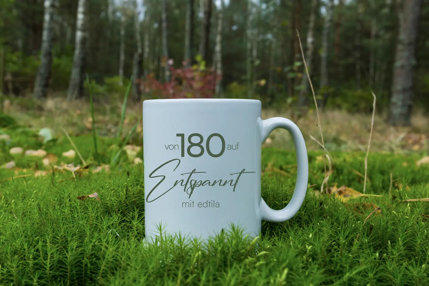 Weiße Keramiktasse mit der Aufschrift „von 180 auf Entspannt – mit edtila“ steht auf weichem, moosbedecktem Waldboden. Im Hintergrund ist ein ruhiger Mischwald mit Birkenstämmen und herbstlichen Farben zu sehen. Die Szene vermittelt Gelassenheit, Naturverbundenheit und innere Ruhe – passend zum Thema Stressabbau und mentale Entspannung.